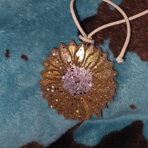 May include: A sunflower-shaped ornament with a glittery design. The petals are gold and brown, with a pink and silver center. A white cord is attached for hanging. The ornament is likely made of wood or a similar material.