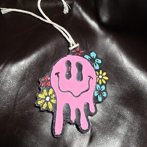 May include: A pink, dripping smiley face air freshener with colorful flower accents. The air freshener has a white string for hanging. The design is on a black background.