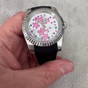 Peut inclure: Montre argentée avec bracelet noir. Le cadran blanc est orné de motifs floraux roses et d'accents bleus. La montre a une lunette texturée et une couronne. Un accessoire élégant pour le poignet.