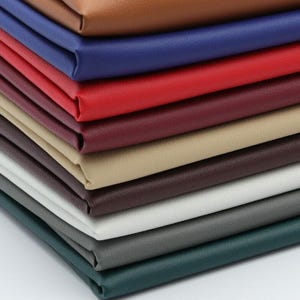 May include: A stack of colorful leather swatches in various shades, including brown, blue, red, burgundy, beige, and gray. The leather appears smooth and has a slight sheen, suggesting a high-quality material. The arrangement showcases the range of colors available.