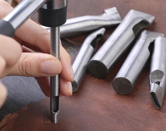 Puede incluir: Primer plano de herramientas de marroquinería. Se utiliza un punzón de metal para crear un agujero en cuero marrón. Se ven varios otros punzones de metal en el fondo.
