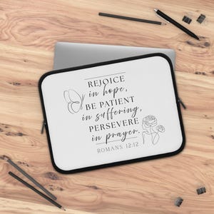 May include: A white laptop sleeve with a black border featuring the text "REJOICE in hope, BE PATIENT in suffering, PERSEVERE in prayer. ROMANS 12:12" in black script. The sleeve has a butterfly and floral design.