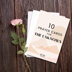 May include: A stack of 10 prayer cards for the unknown, with a soft peach and beige design, and the text "10 Prayer Cards for The Unknown". A pink rose and green leaves are on a dark wood surface.