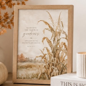 May include: A rectangular light wood frame containing a print of a dried corn stalk in a field. The text reads: HE CROWNS THE YEAR WITH goodness AND YOUR PATHS DRIP WITH ABUNDANCE. PSALM 65:11. The artwork features muted brown, tan, and orange tones.