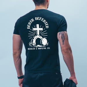 Op de afbeelding: Donkergrijs t-shirt met een witte afbeelding op de rug. De afbeelding bevat een kruis, de woorden "DEATH DEFEATED" en "HEALED & HOPEFUL CO."