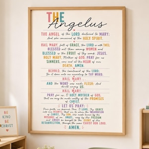 May include: A framed wall art print titled The Angelus featuring the full prayer text in a multi-coloured sans-serif and script font. The design includes small heart icons between lines of text on a cream background, encased in a light wood frame.