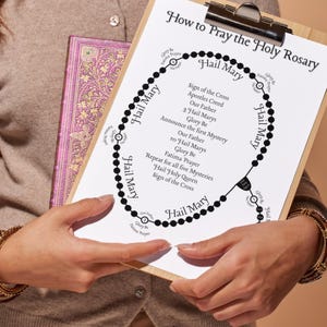 May include: A wooden clipboard holding a white instructional sheet titled How to Pray the Holy Rosary. The paper features a black circular diagram of rosary beads with text labels for prayers, including Hail Mary, Our Father, and Glory Be.