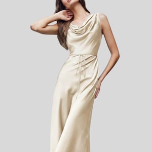 May include: A sleeveless midi-length dress in a champagne-colored satin fabric. The garment features a draped cowl neckline, a fitted bodice, and a thin matching tie belt at the waist. The skirt flows into a straight silhouette that falls below the knee.