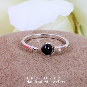 Peut inclure: Bague argentée avec une pierre noire sphérique et une petite pierre d'accent claire. Le design ouvert est mis en valeur sur un fond neutre. Le texte "FRSTORE 26 Handcrafted Jewellery" est visible en bas.