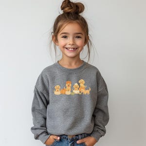 May include: A gray sweatshirt featuring a graphic of several golden retriever puppies. The puppies are depicted with pumpkins and fall-themed accessories. The sweatshirt is worn by a young girl with her hair up in a bun.