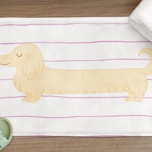May include: A white bath mat with a light yellow dachshund illustration and pink horizontal stripes. A folded white towel and a pair of light green sandals are also visible. The mat is rectangular.