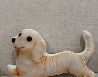 May include: A plush, light tan dog-shaped pillow with black eyes and a smiling expression. The pillow is lying down, with its tail curved upwards. The dog's ears and body have darker tan shading.