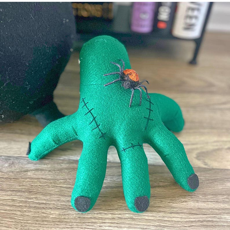 May include: A green felt hand with black stitching and black fingertips. A small, orange and black spider is attached to the hand. The hand is a Halloween decoration.