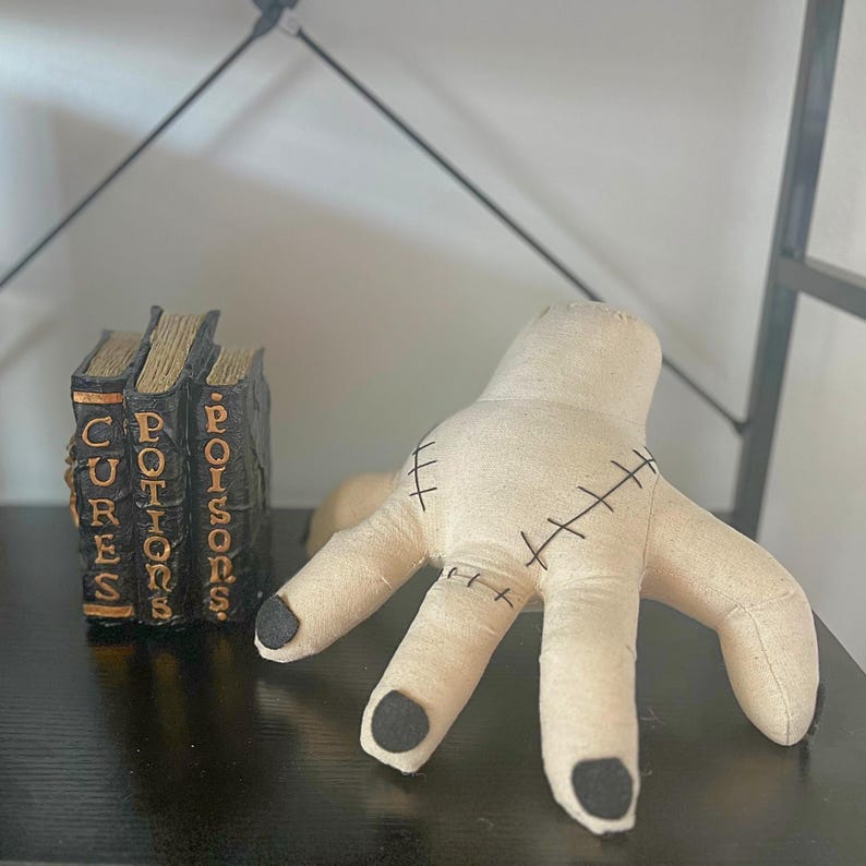 May include: A beige fabric hand with black stitched details and black fingertips. Beside it are three small, black books with gold lettering that reads "CURES", "POTIONS", and "POISONS". The items are displayed on a black shelf.
