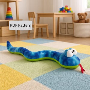 Puede incluir: Un peluche de serpiente azul y verde con grandes ojos blancos y una lengua roja. La serpiente está sobre una alfombra colorida. La imagen incluye el texto "PDF Pattern".