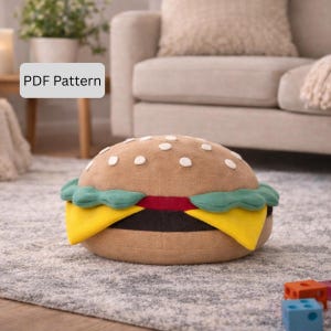 Könnte beinhalten: Ein kissenförmiger Burger mit einem braunen Brötchen, weißen Sesamsamen, grünem Salat, gelbem Käse und einer roten Tomatenscheibe. Das Kissen liegt auf einem grauen Teppich. Der Text "PDF Pattern" befindet sich in einem weißen Feld.