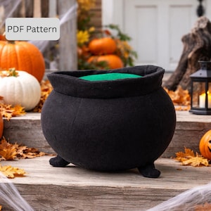 May include: A black felt cauldron with three small feet, a wide rim, and a green interior. The cauldron is a Halloween decoration, sitting on a wooden surface with pumpkins and autumn leaves in the background. Text in the upper left corner reads "PDF Pattern."