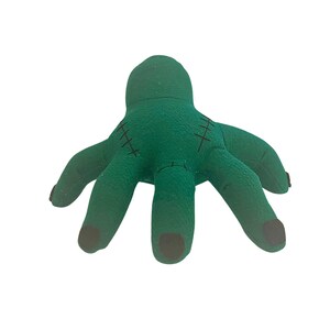 May include: A plush green hand with black stitched details and black fingertips. The toy hand has a Frankenstein monster theme, with a soft, textured appearance. The fingers are spread out, giving the hand a playful look.