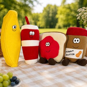 May include: A set of four plush toys shaped like food items: a yellow banana, a red and white striped drink cup with a straw, a slice of bread with red jam, and a brown peanut butter jar with a label reading PEANUT BUTTER and a peanut graphic.