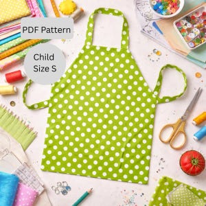 Puede incluir: Un delantal verde con lunares blancos para niños, talla S. El patrón es PDF. Tiene lazo para el cuello y cintas para la cintura. Suministros de costura, como tijeras, hilo y botones, están dispersos.