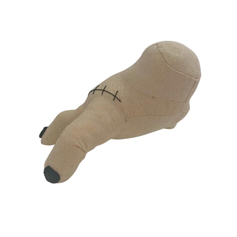 May include: A beige fabric plush toy shaped like a hand, with a thumb and two fingers. The hand has black stitching and dark gray painted fingernails. The toy is made of a soft, textured material.
