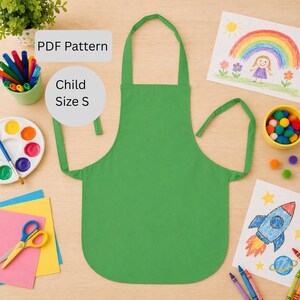 May include: A bright green child-sized apron with the text "PDF Pattern" and "Child Size S". The apron is surrounded by art supplies, including crayons, paint, and drawings of a rainbow and a rocket ship.