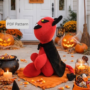 May include: A plush red devil doll with black horns, a black cape, and yellow eyes. The doll is sitting on an orange mat, surrounded by Halloween decorations, including pumpkins and candy corn. The text "PDF Pattern" is visible.