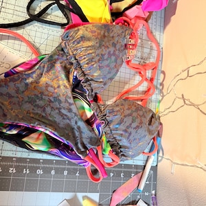 May include: A two-piece swimsuit with a metallic, textured pattern in shades of gray and gold. The swimsuit has bright pink straps and is lying on a cutting mat with a pattern piece and other fabric scraps.