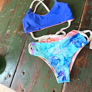 May include: A two-piece swimsuit set. The top is solid blue with white straps and a coral lining. The bottoms feature a watercolor design in shades of blue, pink, and purple, with white straps and a coral lining. The set is displayed on a weathered wooden surface.