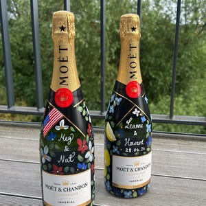 May include: Two Moët & Chandon Imperial Brut champagne bottles with custom hand-painted floral designs. One bottle features the names Meg & Nat with a US flag, while the other displays Leanne & Howard 28.06.26 with lemon accents. Both have gold foil.