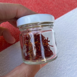 Könnte beinhalten: Ein kleines, klares Glas mit weißem Deckel, gefüllt mit getrockneten Safranfäden. Der Safran hat eine tiefrote Farbe. Das Glas wird in einer Hand gehalten.