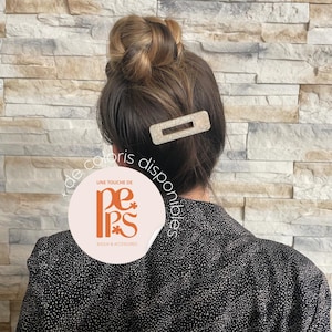 May include: A rectangular, glittery hair clip is worn in the hair. The hair is styled in a bun. The person is wearing a black and white polka dot top. The image has a neutral-toned brick wall background.