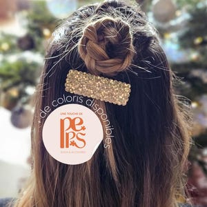 May include: A woman's hair styled with a braided bun and a gold-colored rectangular hair clip. The clip is covered in small, sparkling embellishments. The hair is long and brown, with a blurred background of a Christmas tree.