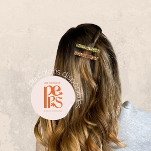 May include: Two sparkly hair barrettes in shades of gold and pink are clipped into wavy brown hair. The hair is styled with a side part. The image includes a logo with the text "Une touche de" and "de coloris disponibles".