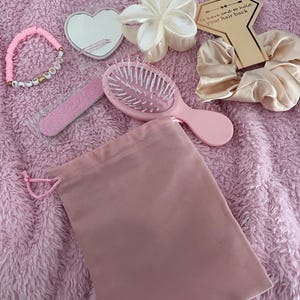 May include: A selection of pink accessories including a bracelet, heart-shaped mirror, flower hair clip, hairbrush, scrunchie, nail file, and a drawstring bag. The bracelet has gold beads spelling out a word. A tag reads "To have and to hold your hair back."