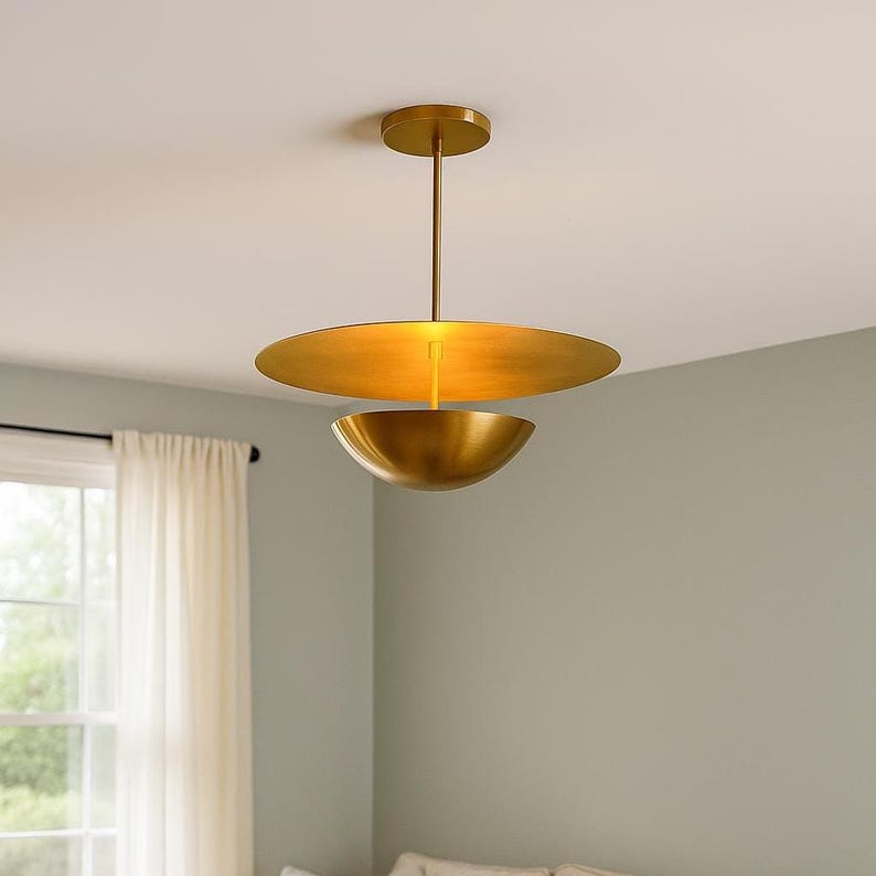 May include: A modern gold-colored pendant light fixture. The light has a circular top piece and a bowl-shaped bottom piece. The fixture hangs from a thin rod attached to a circular ceiling mount. The light is suspended in a room with a window and curtains.