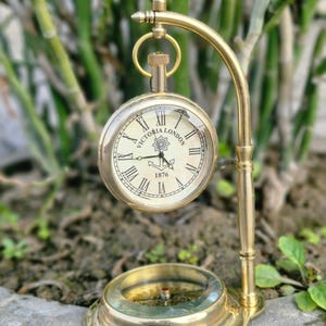 May include: A brass pocket watch and compass set. The watch, marked "Victoria London 1876," features Roman numerals on a cream-colored face. The compass sits below, and the set is mounted on a curved brass stand, showcasing a vintage aesthetic.