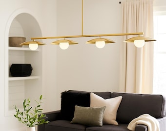 May include: A modern, gold-toned linear chandelier with four lights. Each light features a round, white globe encased in a downward-facing, saucer-shaped shade. The fixture hangs above a dark gray sofa with decorative pillows and a cream-colored throw blanket.