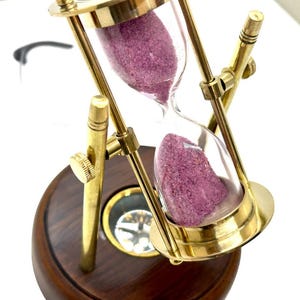 May include: An hourglass with a brass frame and a dark wooden base. The glass chamber contains pink sand. The hourglass is tilted, showing the sand flowing from the top chamber to the bottom. A compass is visible on the base.