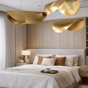 May include: Bedroom interior featuring a bed with white bedding, brown and beige pillows, and a wooden tray with a cup. Above the bed, three large, gold-colored, abstract pendant lights hang from the ceiling.