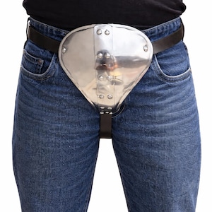 May include: A polished metal codpiece, riveted and attached to a black leather belt and straps. The protective armor is worn over blue denim jeans. The metal piece is shaped to cover the groin area.