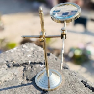 May include: A brass magnifying glass on a stand. The round lens is held by a brass frame and is adjustable. The stand has a circular base and a vertical support with adjustable arms. The magnifying glass is designed for reading or detailed observation.
