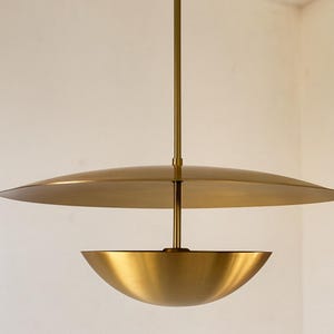 May include: A modern, brass-toned pendant light fixture. The design features a large, flat disc above a bowl-shaped shade, suspended from a central rod. The fixture has a warm, metallic finish, suitable for interior lighting.