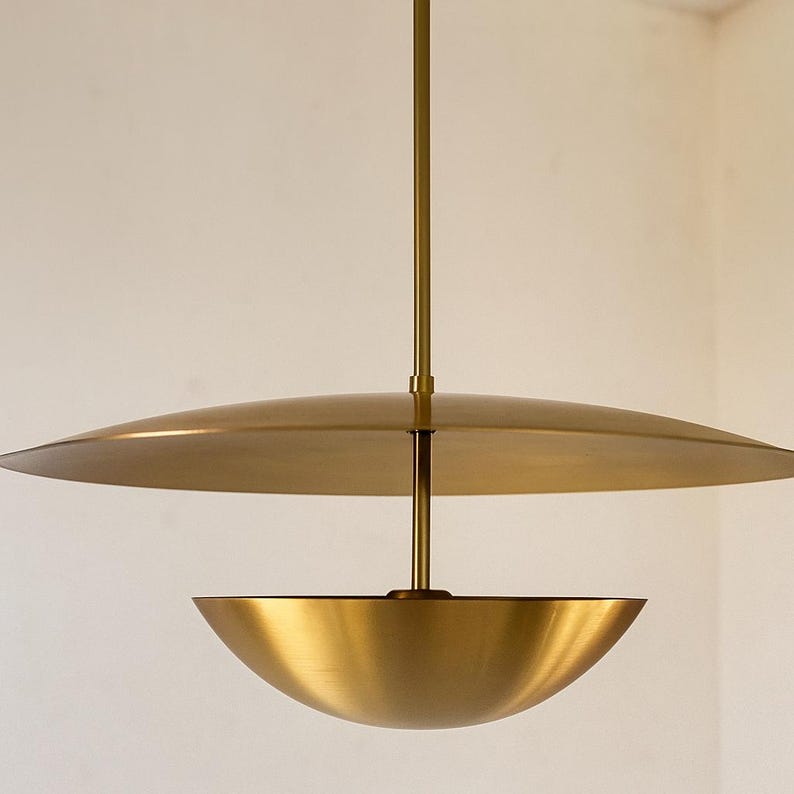 May include: A modern, brass-toned pendant light fixture. The design features a large, flat disc above a bowl-shaped shade, suspended from a central rod. The fixture has a warm, metallic finish, suitable for interior lighting.