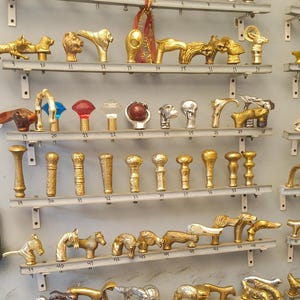 Könnte beinhalten: Eine Sammlung dekorativer Stockgriffe in Gold- und Silbertönen, mit verschiedenen Designs wie Schädeln und Tierfiguren. Die Griffe sind auf Regalen ausgestellt. Einige Griffe haben eine seilartige Textur. Maße nicht verfügbar.