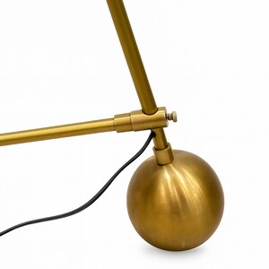 以下が含まれることがあります： 金色のデスクランプのクローズアップ。ベースは球形で、長い調整可能なアームと黒い電源コードが付いています。ランプは金属製で、モダンなデザインです。