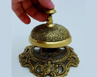 May include: A vintage brass bell with intricate floral and scrollwork detailing. The bell has a dome shape and sits on a decorative base. The bell is being rung by a hand.