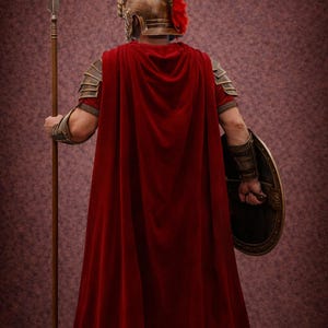 K&ouml;nnte beinhalten: Eine Person in einem r&ouml;mischen Soldatenkost&uuml;m, von hinten gesehen. Das Kost&uuml;m umfasst einen roten Umhang, einen bronzenen Helm mit roter Feder und einen Schild. Der Soldat h&auml;lt einen Speer. Der Hintergrund ist eine strukturierte, kastanienbraune Farbe.