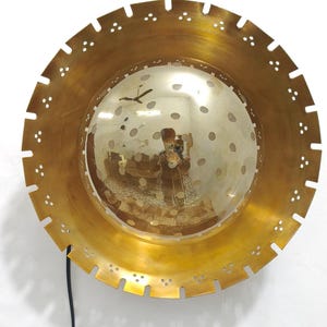 Op de afbeelding: Een messingkleurige, cirkelvormige wandlamp met een reflecterend, bolvormig midden. De buitenrand heeft een reeks uitgesneden vormen en decoratieve perforaties. Een zwart snoer loopt van de onderkant. De lamp is aan een witte muur bevestigd.