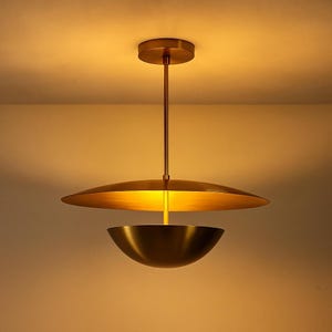 May include: A modern, brass-toned pendant light fixture. The design features a large, flat disc above a bowl-shaped shade, suspended from a ceiling mount by a slender rod. The warm gold color suggests an elegant, contemporary aesthetic.